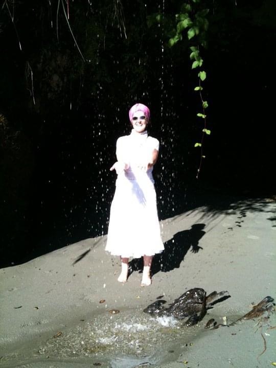 Phoebe standing under a waterfall in the sunlight on the Pacific Coast with arms outstretched.